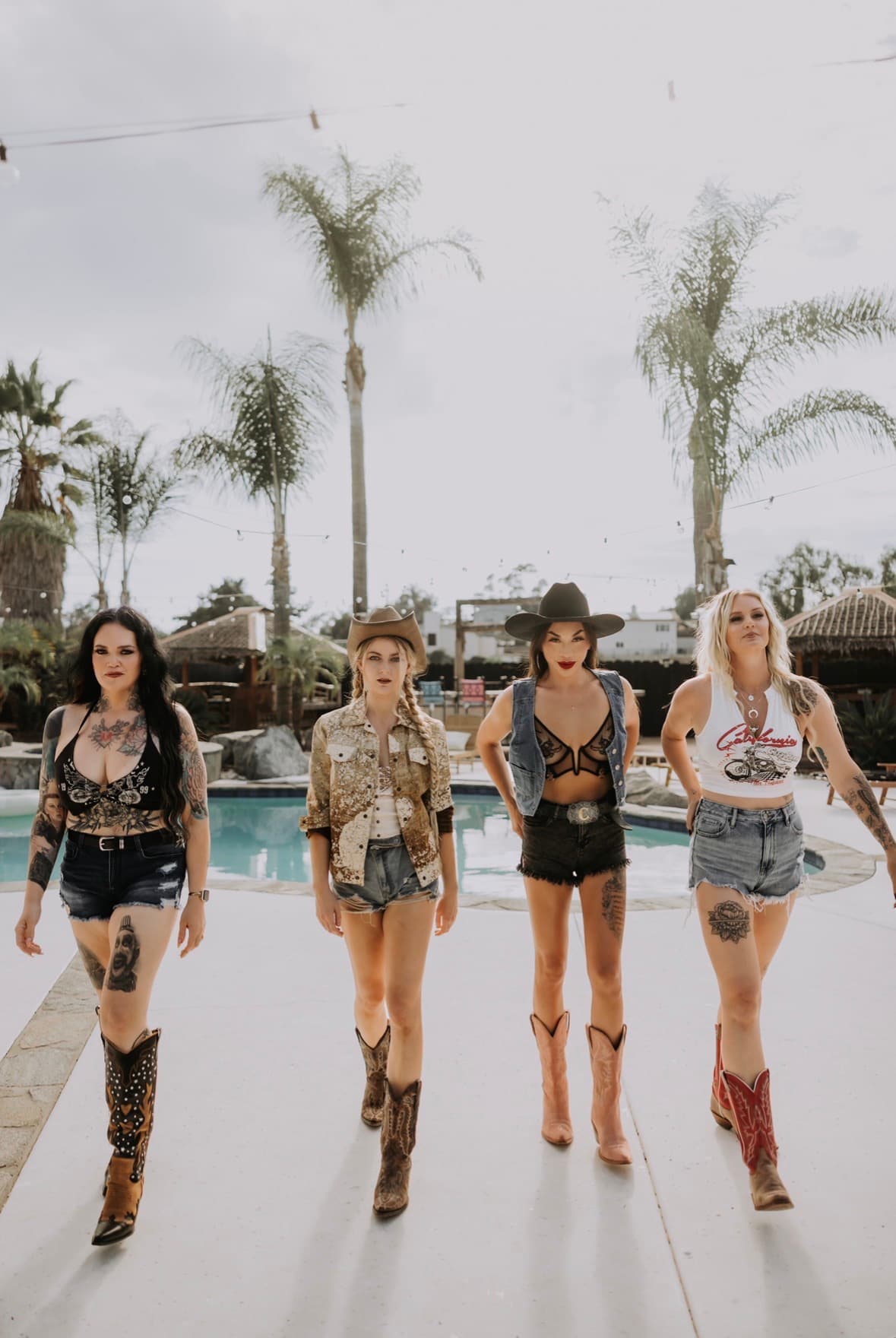 Four women in cowboy boots walking beside a pool under palm trees